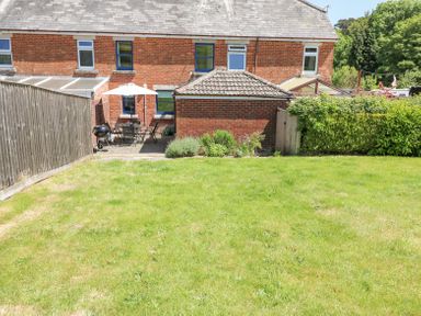A backyard with grass lawn a patio with table chairs umbrella and barbecue next to a brick house and wooden fence at Studland Cottage in Studland