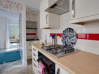 A kitchen with a gas stove a patterned tray and utensils next to an open doorway showing a room with a sofa and leaf patterned wallpaper at Sunny Days in Weymouth