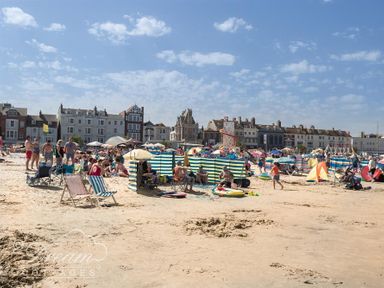 A crowded beach with people seated on chairs and towels with windbreakers and buildings in the background at Sunny Days in Weymouth