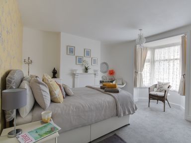 A bedroom with a bed, side table with a lamp and a magazine, an armchair by a bay window with curtains at Sunny Days in Weymouth