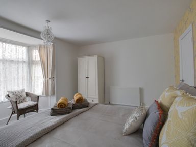 A bedroom with a bed with pillows and folded towels a wicker chair by the window and a white wardrobe at Sunny Days in Weymouth