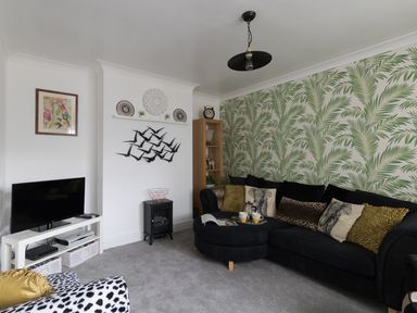 A living room with a black sectional sofa with patterned cushions a wooden shelving unit and a tv on a white stand at Sunny Days in Weymouth