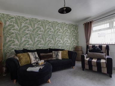 A living room with a black sofa and cushions next to a striped armchair with cushions and a large window at Sunny Days in Weymouth