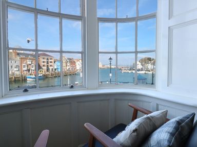 A bay window with armchairs and cushions overlooking a harbor with boats and buildings at Tradewinds Brewers Quay Harbour