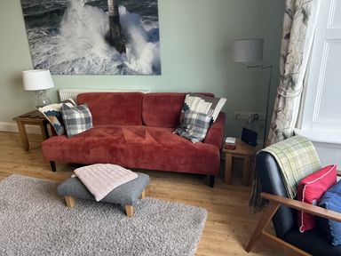 A red sofa with cushions and a gray footstool with a folded blanket in a living room with wooden floor and a large photograph of a lighthouse at Tradewinds Brewers Quay Harbour