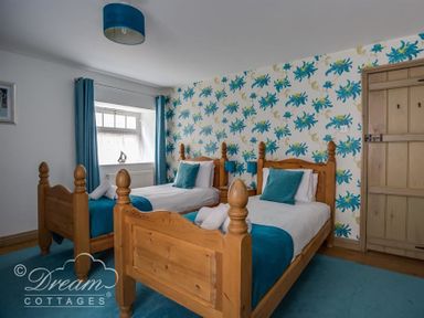A bedroom with two wooden single beds with blue bedding and cushions a floral wallpapered wall a window with blue curtains and a wooden door at Upsidedown House Brewers Quay Harbour