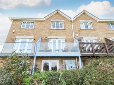 A rear view of a brick house with a glass balcony and patio doors surrounded by bushes at Waters Edge in Wyke Regis