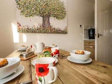 A wooden dining table set with white bowls containing muffins red flower mugs a white teapot and a metal butter dish beneath a painting of a tree at Waters Edge Wyke Regis