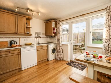 A kitchen with wooden cabinets a dishwasher a washing machine a countertop with bread and a kettle dining table set for two near glass doors to a garden at Waters Edge in Wyke Regis