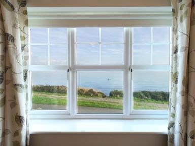 A window with patterned curtains overlooking a coastal path and calm sea at Waters Edge in Wyke Regis