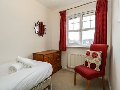 A bedroom with a single bed towels on it a wooden chest of drawers a red chair with a patterned cushion and red curtains at Waters Edge in Wyke Regis