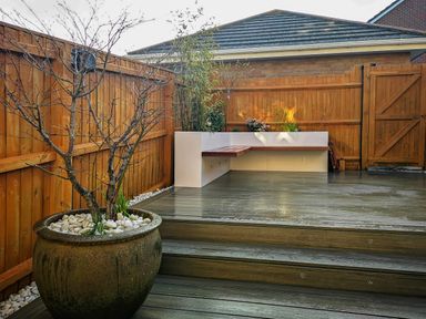 A fenced backyard with wooden decking a large potted tree and a built-in corner bench at Waters Edge in Wyke Regis