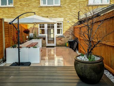 A small backyard patio with a large potted tree a white umbrella a wooden bench and a door to a brick house at Waters Edge in Wyke Regis