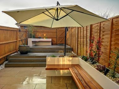 A backyard patio with wooden bench seating under a large umbrella and steps leading to a raised deck with potted plants at Waters Edge in Wyke Regis