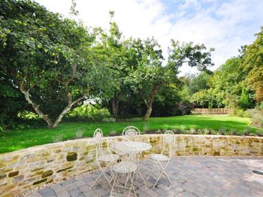 A garden patio with a stone wall and metal table and chairs set surrounded by trees and grass at Warmwell Lodge in Warmwell