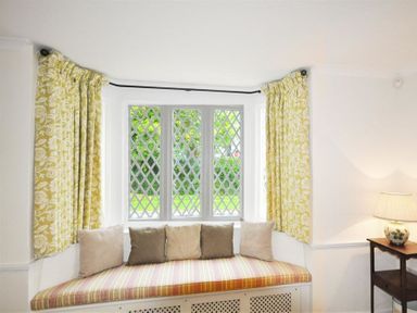 A window seat with striped cushion and five pillows below a diamond pane window with yellow patterned curtains and a side table with lamp at Warmwell Lodge in Warmwell