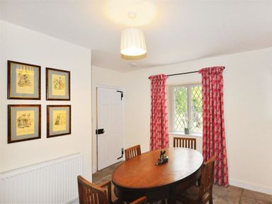 A dining room with a wooden oval table four wooden chairs a window with red patterned curtains and framed pictures on the wall at Warmwell Lodge in Warmwell