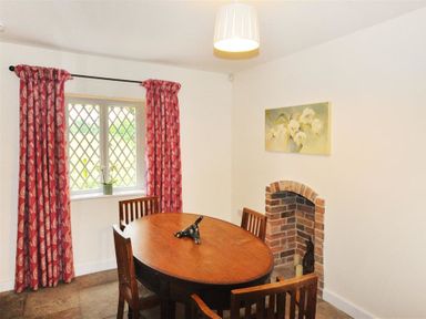 A dining room with a wooden oval table and chairs next to a window with red patterned curtains and a small brick fireplace at Warmwell Lodge in Warmwell