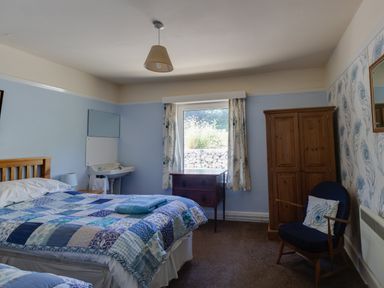 A bedroom with a bed with patchwork quilt a wooden wardrobe a bedside table a sink a window with curtains and a chair at Waterson in Osmington