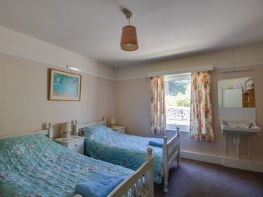 A bedroom with two single beds with blue bedspreads a window with floral curtains a wall-mounted sink and a framed picture on the wall at Waterson in Osmington