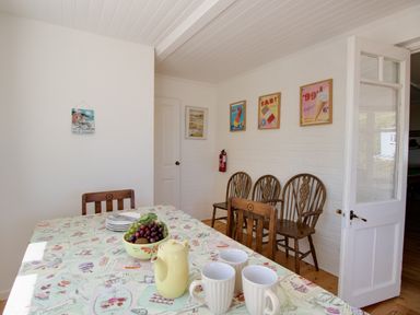 A dining area with a table covered in a patterned tablecloth holding a bowl of fruit teapot and cups wooden chairs against a white brick wall with framed posters at Waterson in Osmington