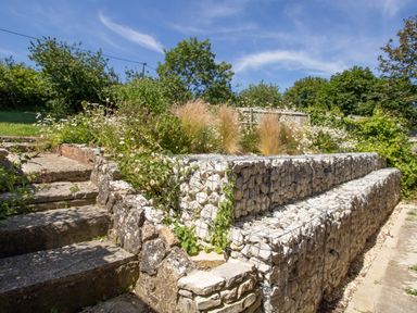 An outdoor garden area with stone steps and a terraced wall made of rocks in wire cages with plants and trees in the background at Waterson in Osmington