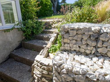 Stone steps next to a house leading up to a garden area with a wire cage filled with rocks and surrounding plants at Waterson in Osmington