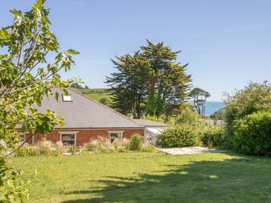 A grassy garden with trees and bushes near a brick house with a sloped roof and a view of the sea at Waterson in Osmington