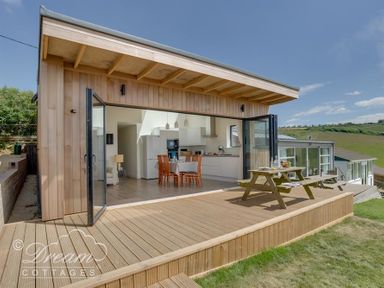 A wooden deck with picnic tables outside a cottage with open glass doors showing a kitchen and dining area at The Wave in Eype