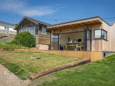 A wooden cottage with open glass doors leading to a dining area and a grassy yard with toys at The Wave in Eype