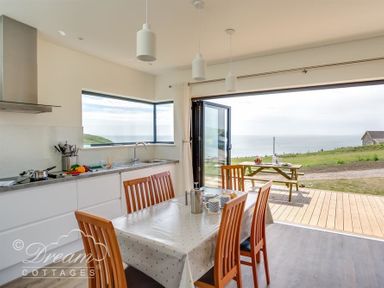 A kitchen and dining area with wooden chairs and table near large open windows overlooking a grassy area and sea at The Wave in Eype