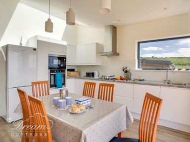 A kitchen with a dining table and wooden chairs next to appliances and a window showing a landscape at The Wave in Eype