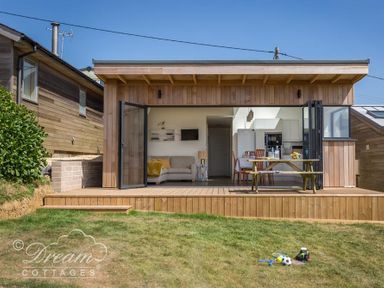 A wooden cabin with a patio table and open glass doors showing a living room and kitchen at The Wave in Eype