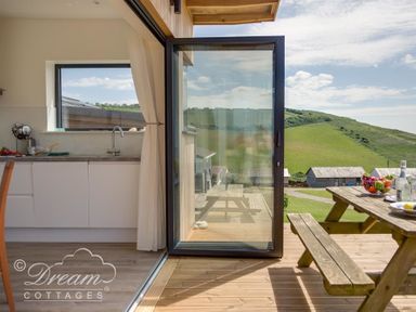 A kitchen with a sink and window opening onto a wooden deck with a picnic table and hills in the background at The Wave in Eype