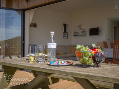 A wooden table with a bottle, glasses, a colorful plate, and a bowl of fruit in a room with a sofa and dining table at The Wave in Eype