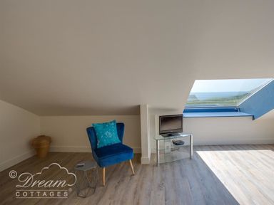 A room with a blue chair, a small side table, a television on a glass stand, and a large skylight window at The Wave in Eype