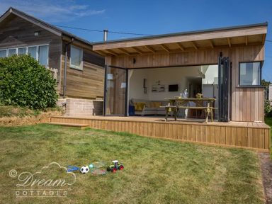 A wooden cottage with open patio doors showing an indoor dining table and living room at The Wave in Eype