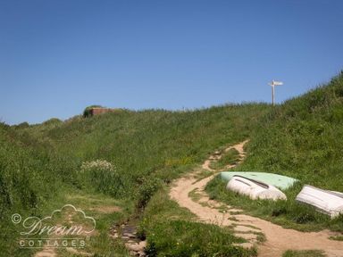 A dirt path winding through grassy hills with three overturned boats on the side and a signpost at The Wave in Eype