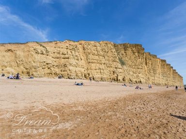 A sandy beach with people sitting and walking near tall yellow cliffs under a blue sky at The Wave in Eype