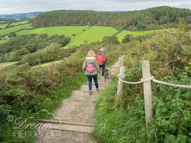 Two people walking down a dirt path with wooden steps and rope railing in a green rural area at The Wave in Eype