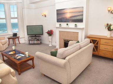 A living room with beige sofas a wooden coffee table a TV near windows showing the sea a wooden sideboard and a fireplace at Weymouth Bay Apartment B in Greenhill