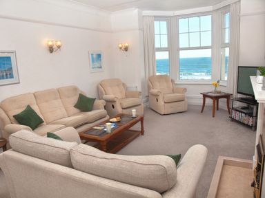A living room with beige sofas and armchairs a wooden coffee table a small side table a flat screen television and a large window with a view of the ocean at Weymouth Bay Apartment B in Greenhill