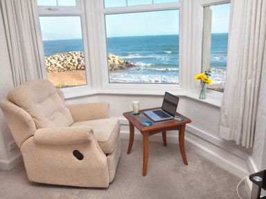 A beige armchair next to a wooden table with a laptop and mug by a window overlooking the sea at Weymouth Bay Apartment B in Greenhill