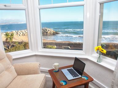A corner seating area with a beige armchair a wooden side table with a laptop and mug and a vase with sunflowers by windows overlooking the sea at Weymouth Bay Apartment B in Greenhill