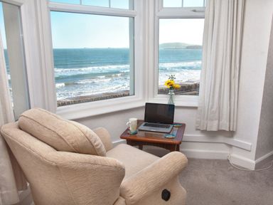 A beige armchair next to a small table with a laptop a mug and a vase with sunflowers by windows overlooking the sea at Weymouth Bay Apartment B in Greenhill