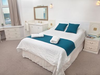 A bedroom with a double bed with blue cushions and a blue runner a bedside table with a magazine and a mug a window showing the sea and a vanity with a mirror at Weymouth Bay Apartment B in Greenhill