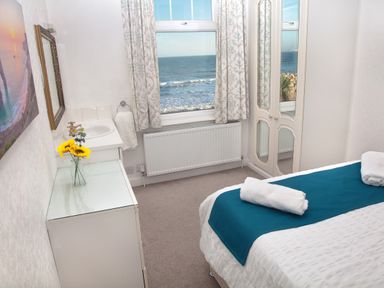 A bedroom with a bed with towels on it a dresser with a vase of flowers a window with curtains and a mirrored wardrobe at Weymouth Bay Apartment B in Greenhill