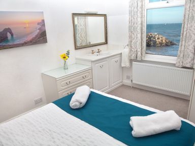 A bedroom with a bed with white and teal bedding a chest of drawers a sink with a mirror a window with curtains overlooking the sea at Weymouth Bay Apartment B in Greenhill