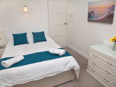 A bedroom with a bed with white and teal bedding two teal pillows a dresser with a vase of sunflowers and a wall picture at Weymouth Bay Apartment B in Greenhill
