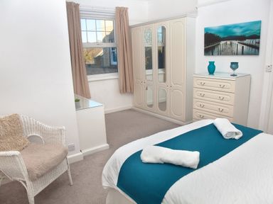 A bedroom with a bed with blue runner and towels a white wardrobe a chest of drawers with vases a wicker chair and a window at Weymouth Bay Apartment B in Greenhill